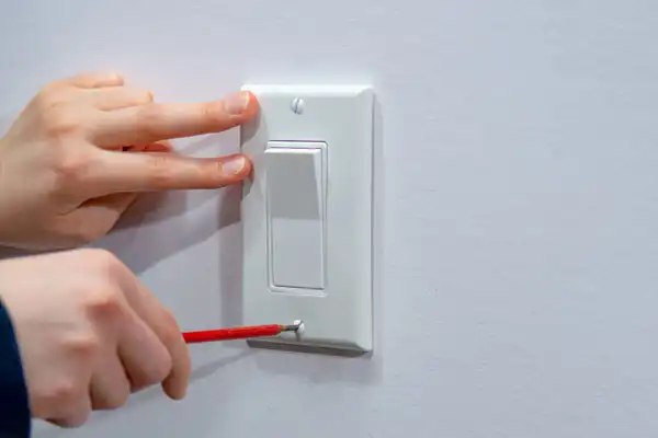 A person uses a red screwdriver to install or adjust a white wall light switch, with another hand steadying the switch plate.